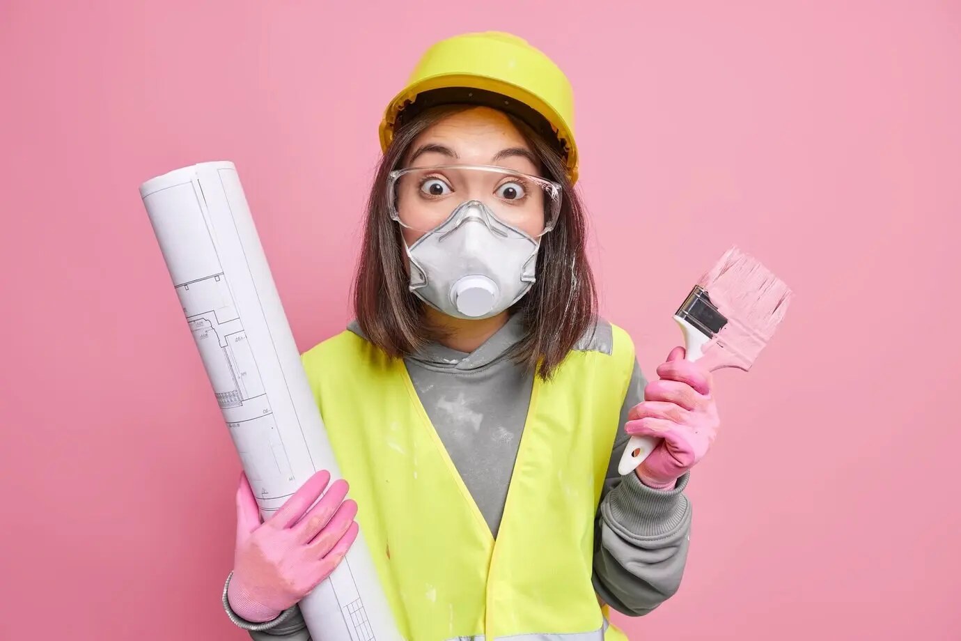 Eine Frau trägt Schutzbrille, Atemschutzmaske und Schutzhelm, hält einen Bauplan und einen Malerpinsel, will die Wände des Hauses neu streichen und posiert vor pinkem Hintergrund.