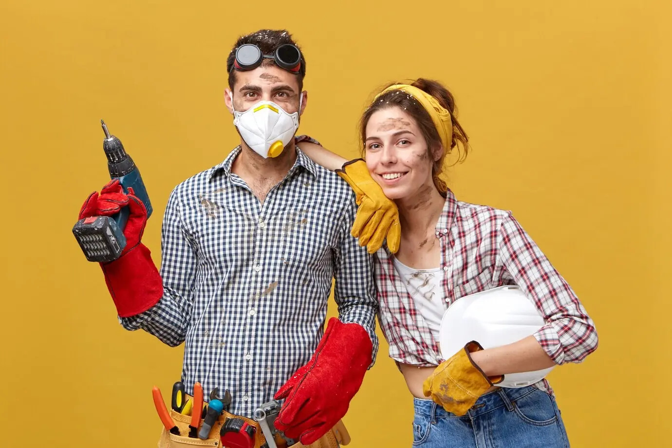 Fröhliches Paar mit schmutzigen Gesichtern in Freizeitkleidung, hält Bohrmaschine und Schutzhelm und führt Reparaturen in ihrem Haus durch. Junger, attraktiver, männlicher Hauseigentümer mit Schutzhandschuhen und Maske bohrt etwas in die Wand.